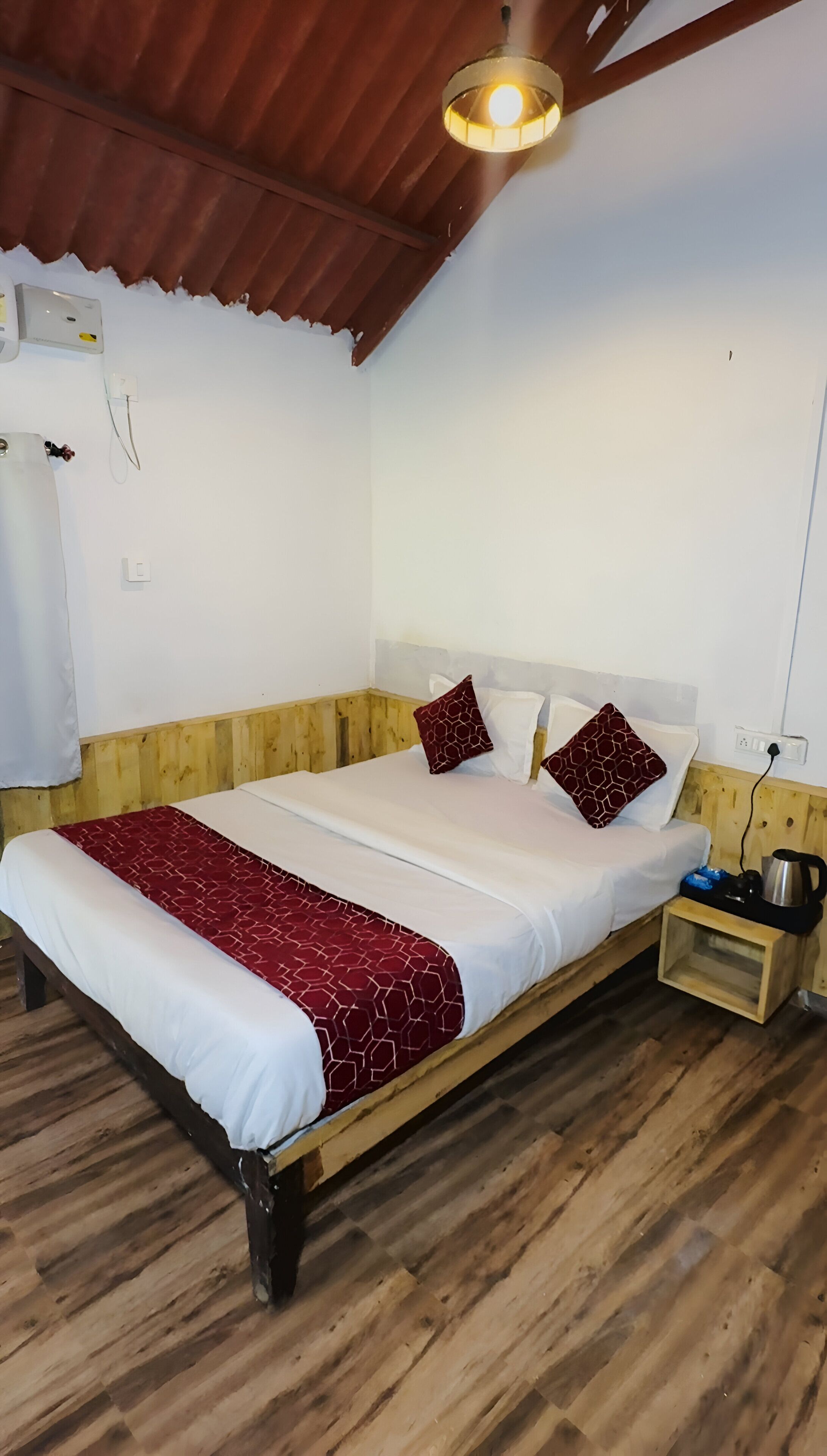 Ein ordentlich gemachtes Bett mit einer roten und weißen Bettdecke, zwei passende Kissen und einem hölzernen Kopfteil. Ein Nachttisch mit Wasserkocher und Tasse, sowie eine Deckenleuchte über dem Bett.