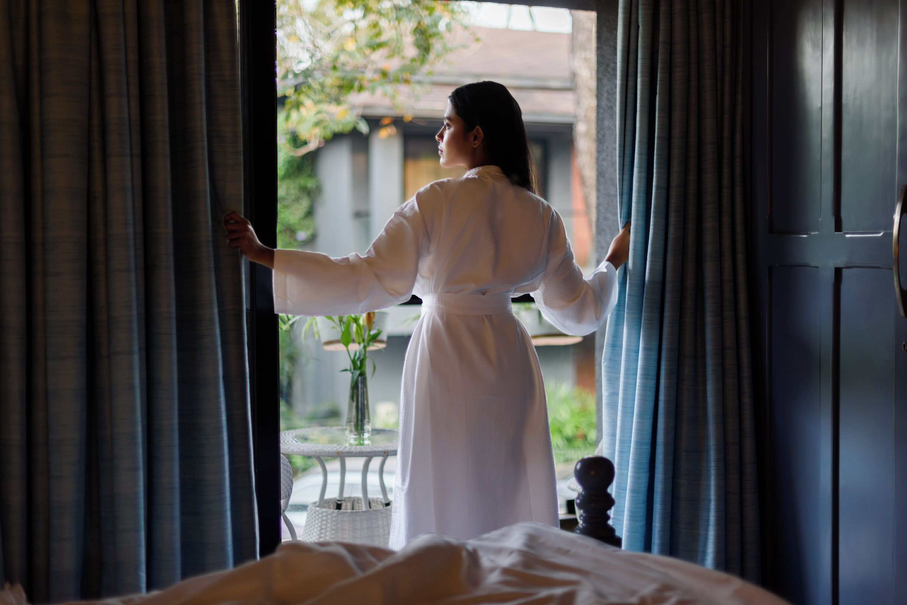 Une personne vêtue d’une robe blanche se tient près d’une fenêtre ornée de rideaux, à côté d’un lit et d’une petite table sur laquelle se trouve un vase de fleurs.