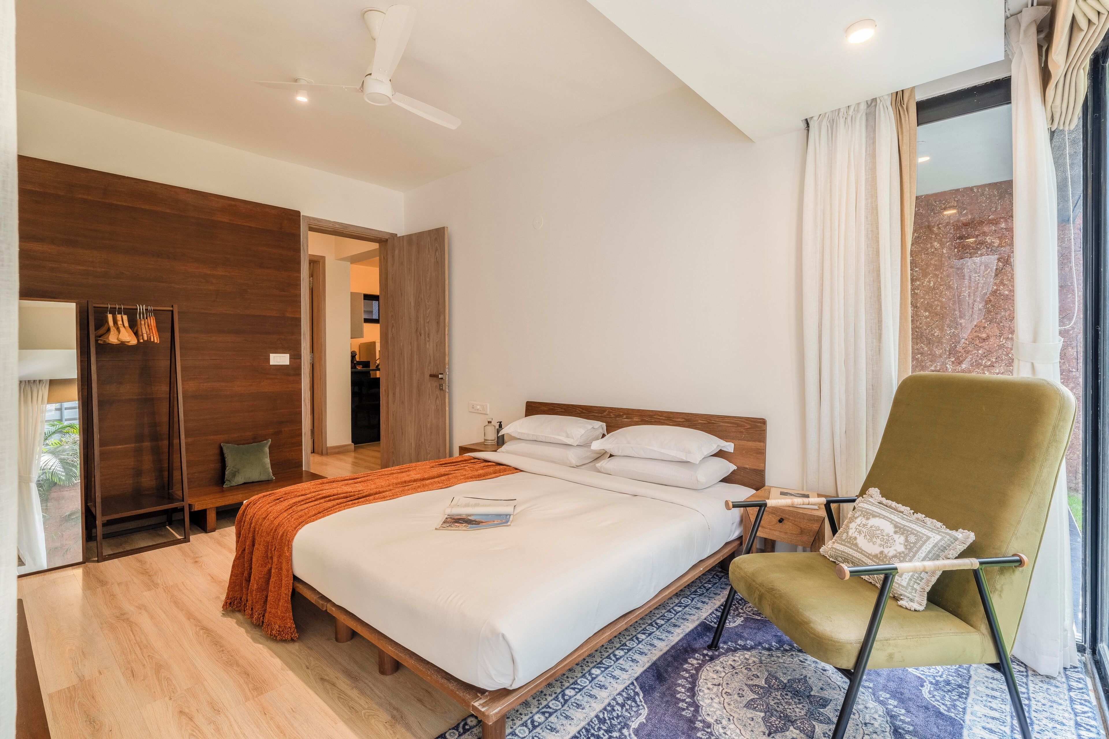 A modern hotel room with a large bed, a wooden wardrobe, a sitting area with a chair, and a view through a window.
