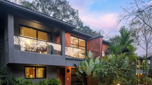 Un bâtiment moderne à deux étages, doté de grandes fenêtres, d’un balcon et d’un jardin bien entretenu, agrémenté de diverses plantes et d’un sentier.