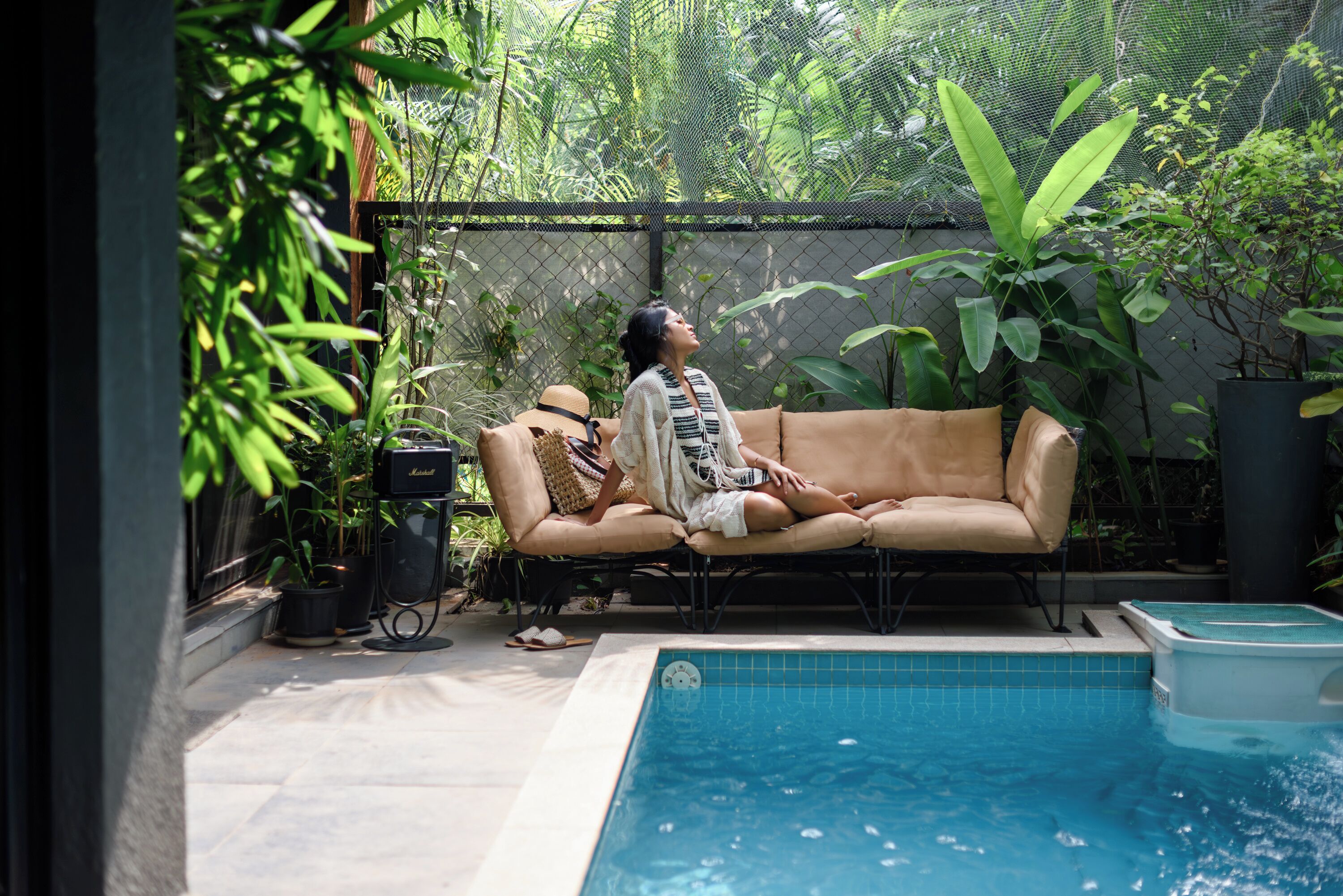 A person lounging on a beige sofa by a pool, surrounded by lush greenery and a fence.