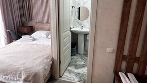 A hotel room with a bed, a bedside table, a chair, a bathroom with a sink and mirror, and a wooden door.