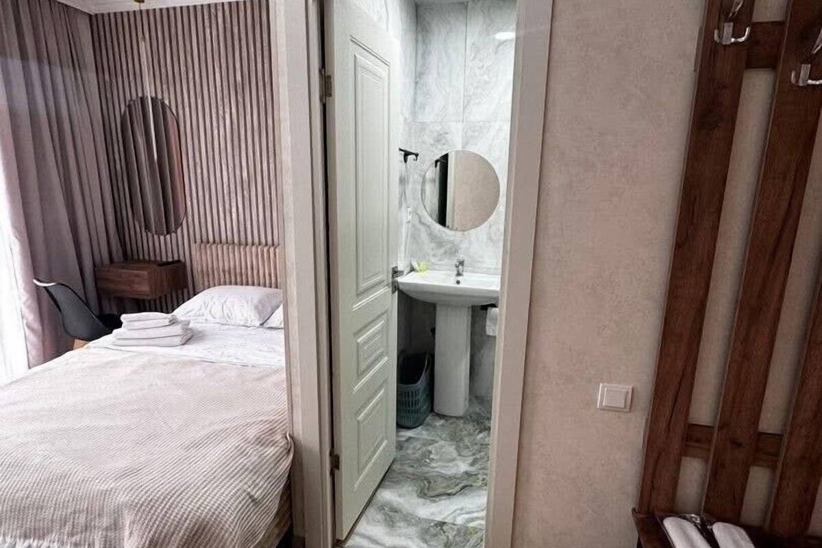 Habitación de hotel con cama, mesita de noche, una silla, baño con lavamanos y espejo, y puerta de madera.