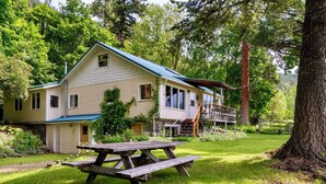 A two-story house with a blue roof, a front porch, and a wooden deck. - A serene, rustic farmhouse nestled on the banks of the Jocko River. Perfect for nature lovers, this cozy getaway offers peace, privacy, and stunning outdoor spaces to immerse yourself in the beauty of Montana. (Arlee)