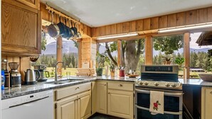 A kitchen with wooden cabinets, a stove, and a large window overlooking a mountainous landscape. - A serene, rustic farmhouse nestled on the banks of the Jocko River. Perfect for nature lovers, this cozy getaway offers peace, privacy, and stunning outdoor spaces to immerse yourself in the beauty of Montana. (Arlee)