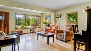A spacious living room with a piano, a dining area with a table and chairs, and large windows offering a view of greenery. - A serene, rustic farmhouse nestled on the banks of the Jocko River. Perfect for nature lovers, this cozy getaway offers peace, privacy, and stunning outdoor spaces to immerse yourself in the beauty of Montana. (Arlee)