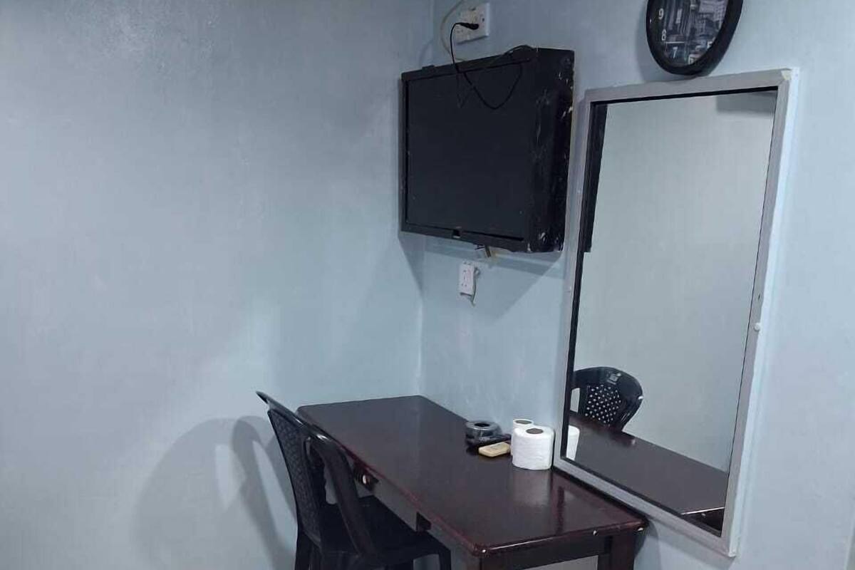 A small room with a wooden table, a black chair, a mirror, a clock, and a trash bin.
