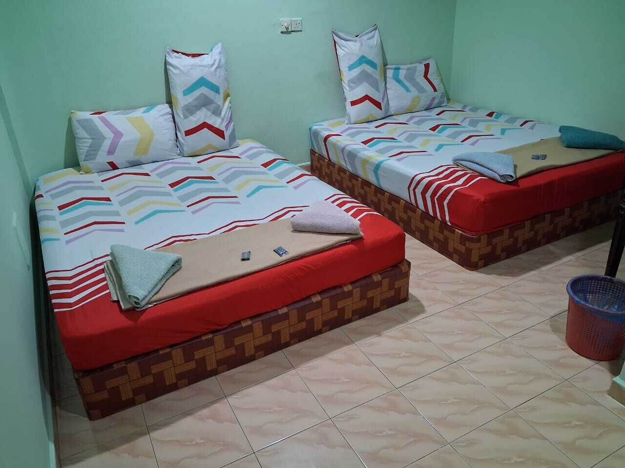 Habitación con dos camas, cada una con ropa de cama y almohadas estampadas. Se ve un bote de basura rojo y una toma de corriente en la pared.