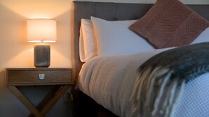 Ein ordentlich bezogenes Bett mit grauem Kopfteil, einer Nachttischlampe und einem Holfnachtkästchen.