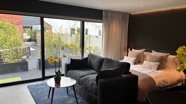 Ett modernt hotellrum med en stor skjutdörr i glas som leder ut till en innergård med växter och en mur av tegelsten.