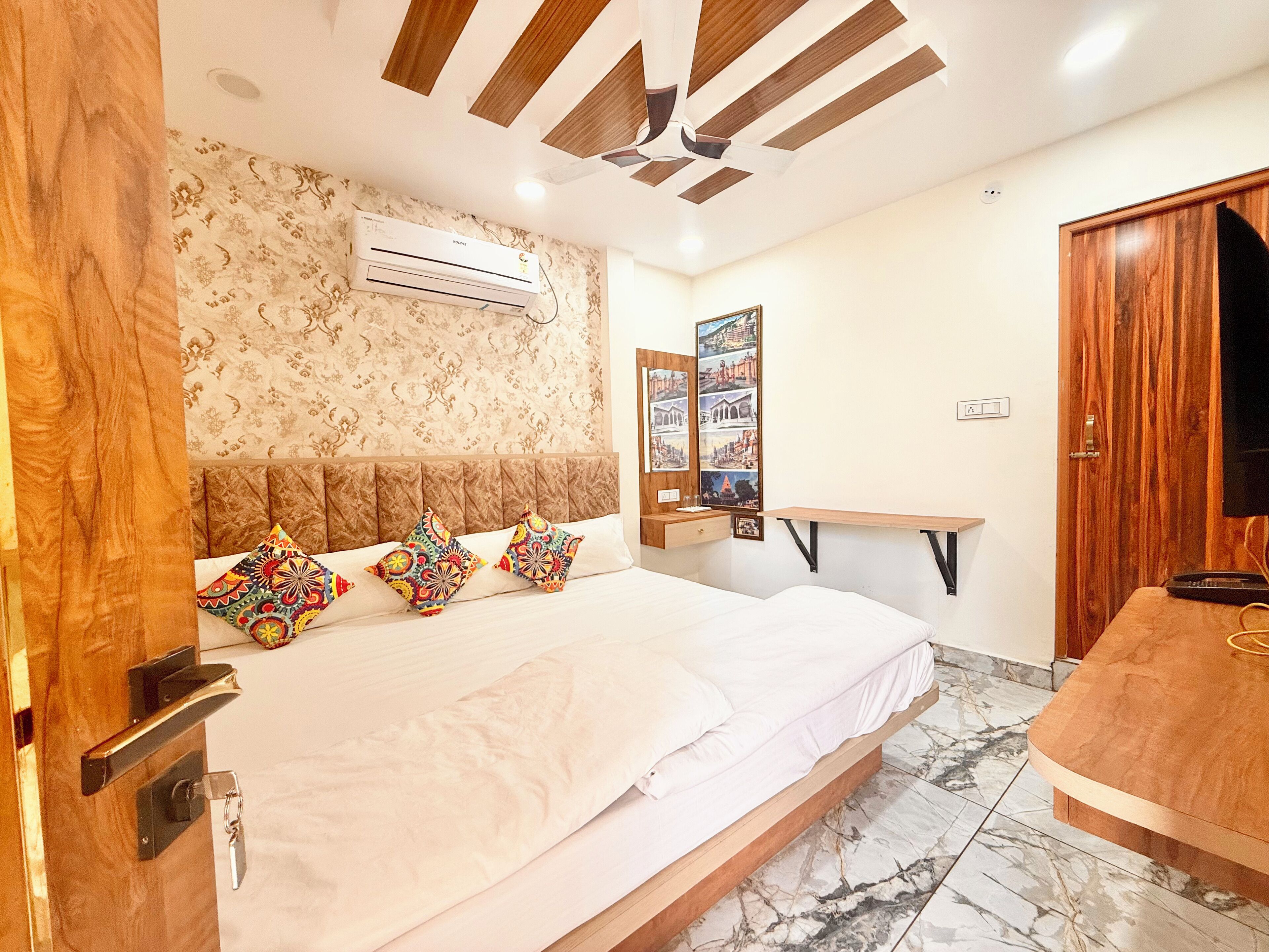 Un dormitorio moderno con una cama grande, un cabecero de madera, un ventilador de techo, un equipo de aire acondicionado, un escritorio de madera y un televisor.