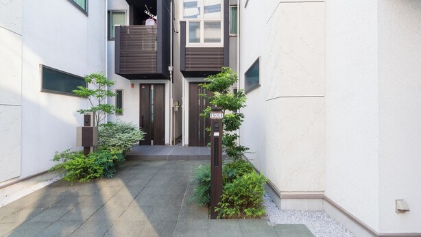 Un estrecho callejón con un pequeño jardín, un buzón y una puerta que da a un edificio de diseño moderno.