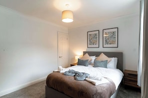A neatly made bed with a brown blanket, a bedside table with a lamp, and two framed artworks on the wall.