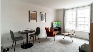 A living room with a round table, two chairs, a television, and a window with curtains.