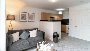 A living room with a sofa, a lamp, framed artwork, a trash bin, and a kitchen area with wooden cabinets and a washing machine.
