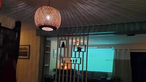 Una habitación con poca luz, una pantalla grande mostrando una página web, un lámpara colgante y una separador de madera con estantes.