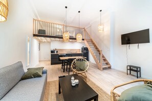 Ein modernes Wohnzimmer mit einer Couch, einem Couchtisch und einem Korbstuhl. Eine Treppe führt zu einer oberen Ebene mit einer Küche und einem an der Wand befestigten Fernseher.