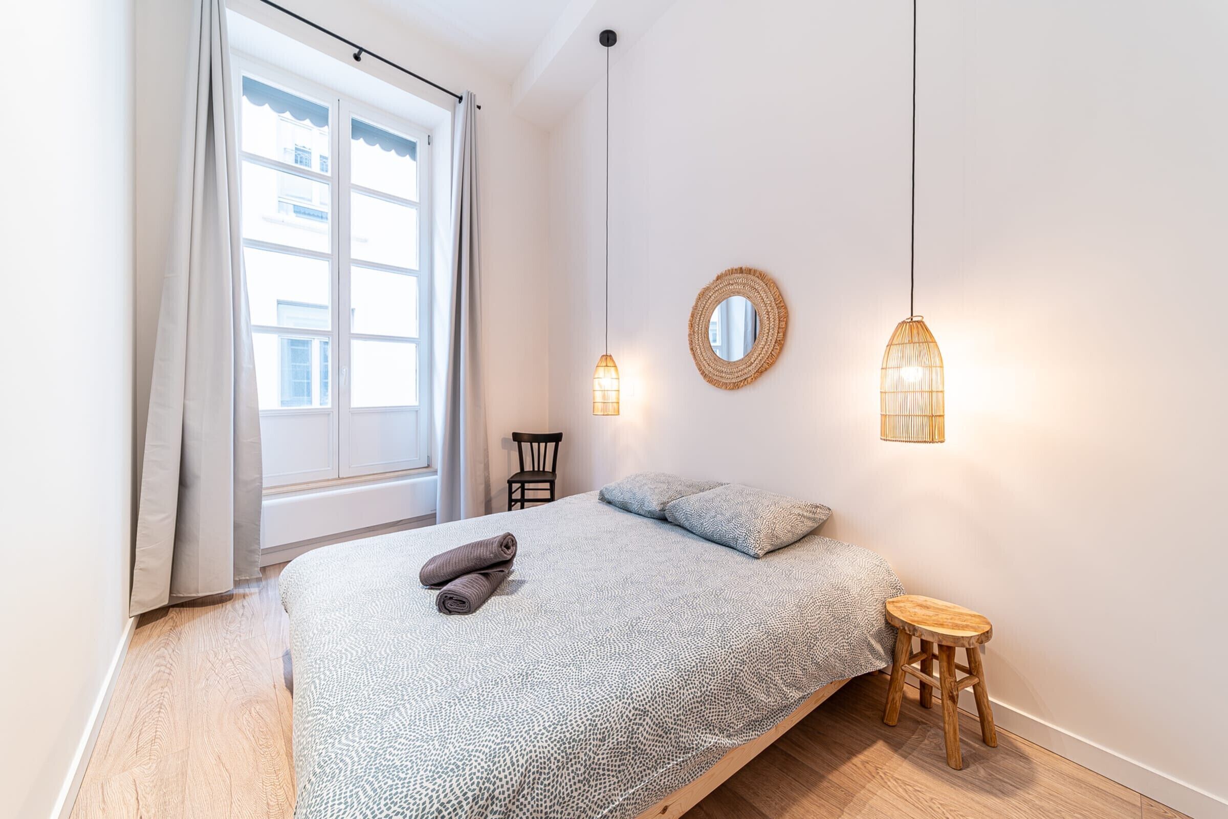 Ein Schlafzimmer mit einem Bett, zwei Pendelleuchten, einem runden Spiegel und einem hölzernen Hocker.