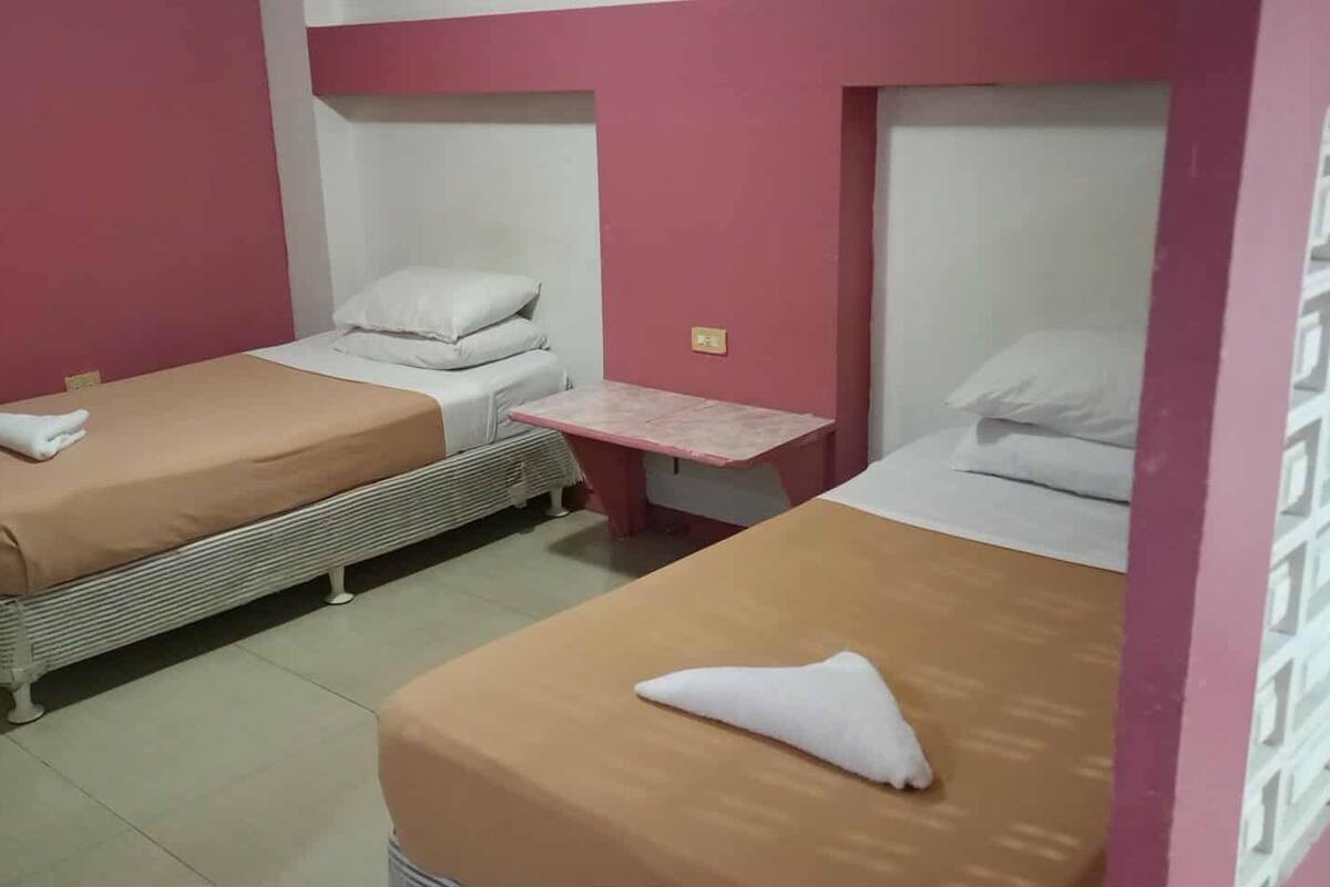 A room with two single beds, each with a pillow and a blanket, a small table, and a wall unit with storage.