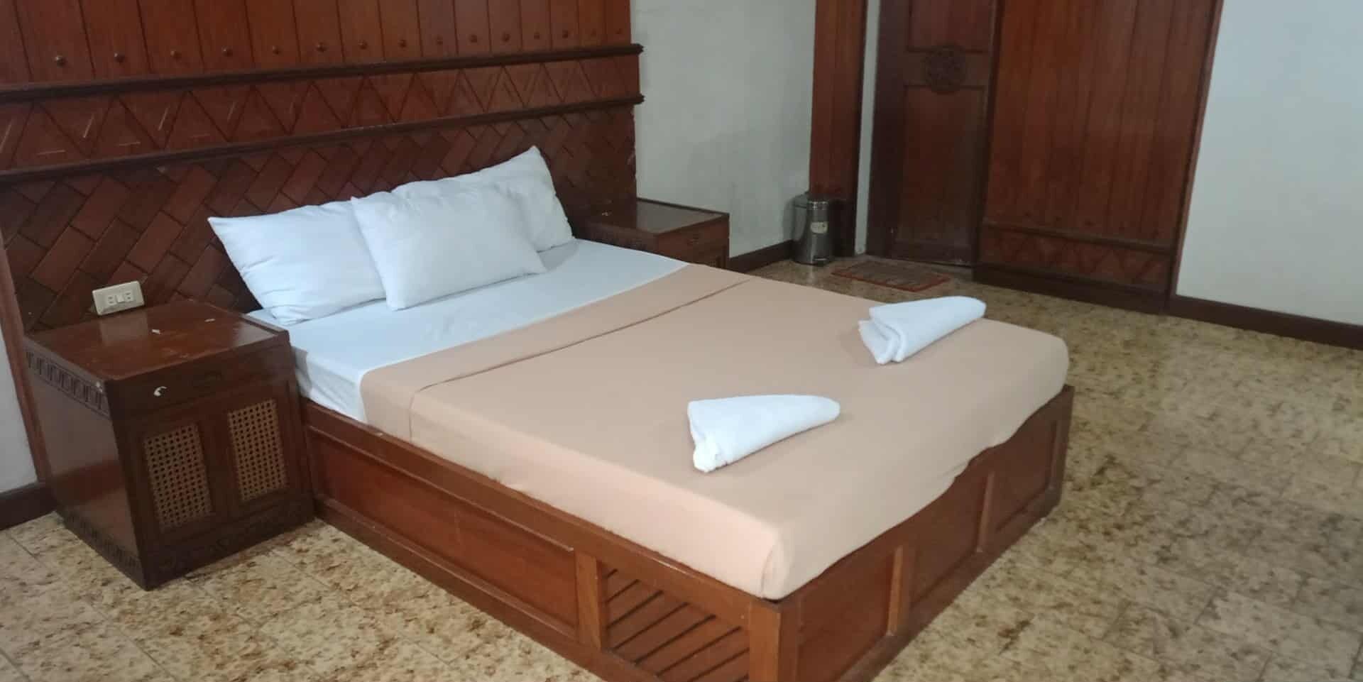 A neatly made bed with a wooden headboard, a beige bedspread, and two folded towels.