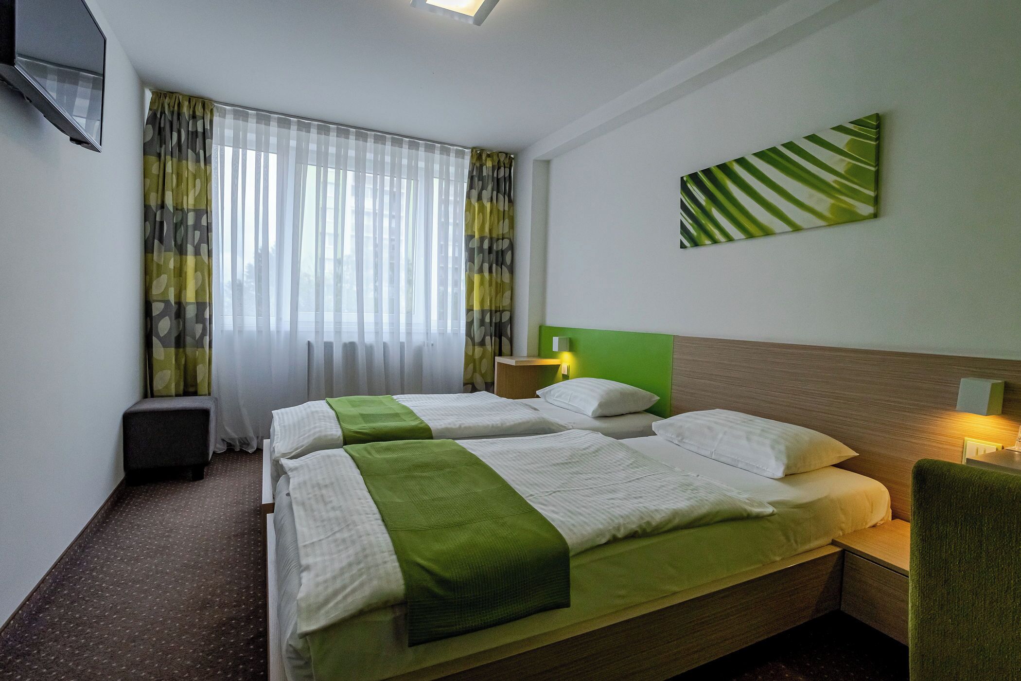 Habitación de hotel con dos camas, cabecero verde, televisor montado en la pared, un cuadro y una ventana con cortinas.