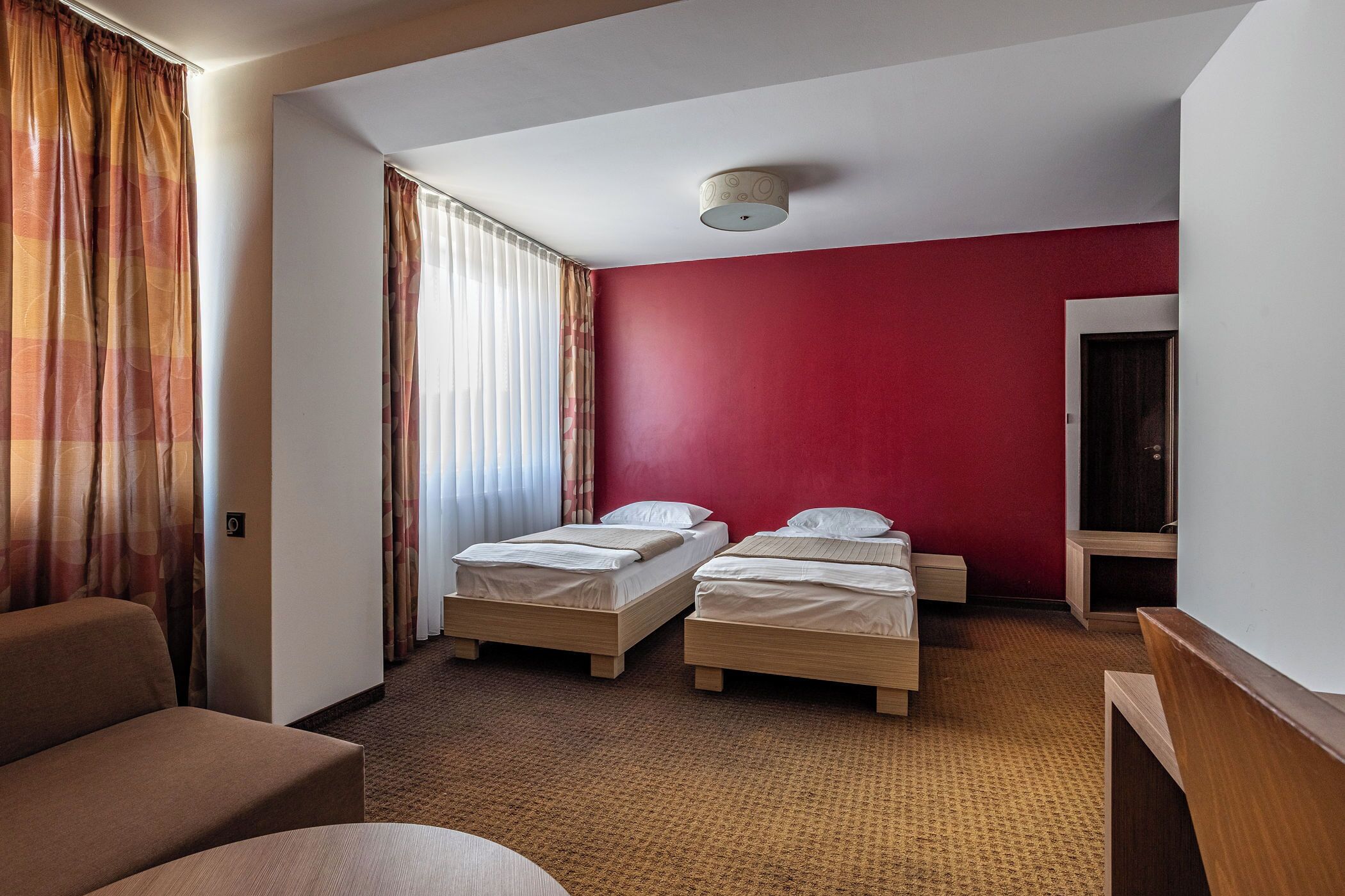 Une chambre d’hôtel avec deux lits simples, un canapé, un bureau en bois et un mur d’accent rouge.