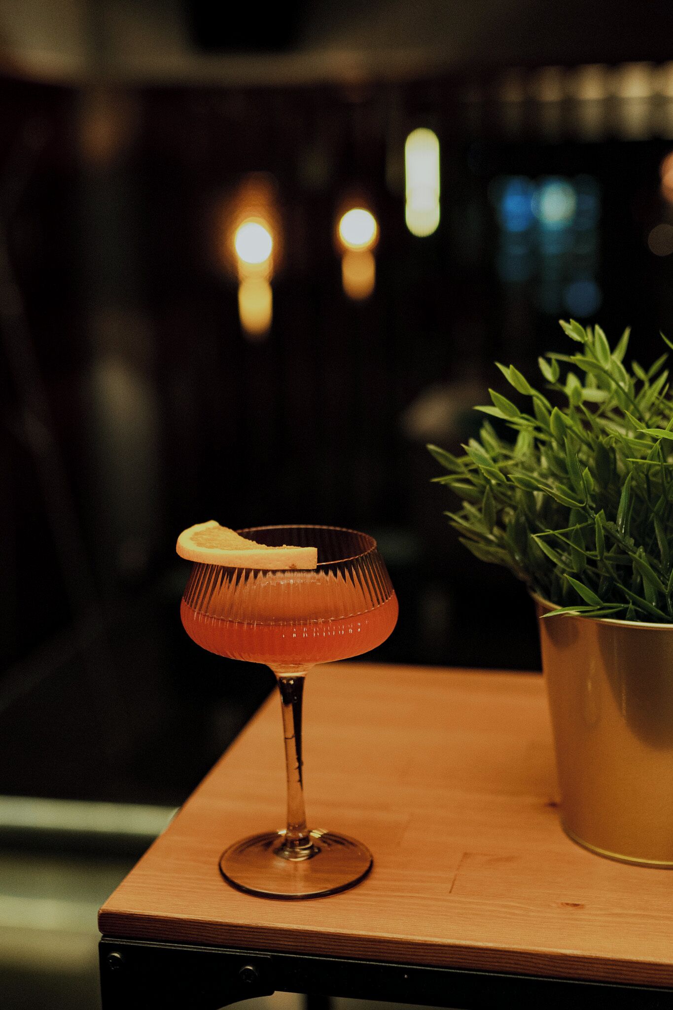 A cocktail glass with a slice of orange on a wooden table, accompanied by a potted plant.