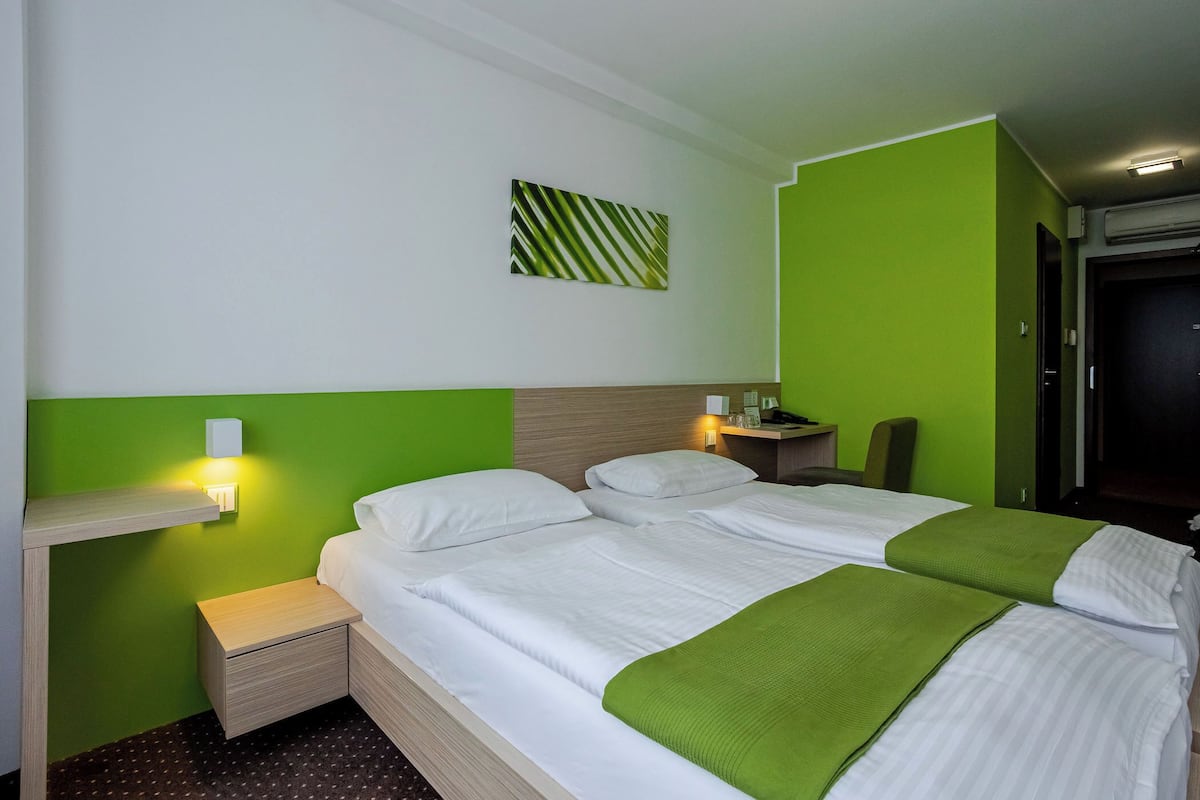 Habitación de hotel con dos camas, una mesita de noche, una lámpara de pared y una pared decorativa verde.