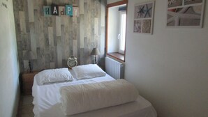 A bedroom with a bed, pillows, a nightstand with a lamp, a wall clock, and a window with a view. - Longère vendéenne (Saint Hilaire de riez)