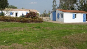Two white buildings with blue shutters, a grassy field, and trees in the background. - Longère vendéenne (Saint Hilaire de riez)