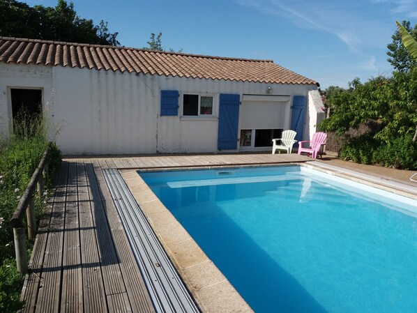 A swimming pool with a wooden deck, a white building with blue shutters, and two white chairs.