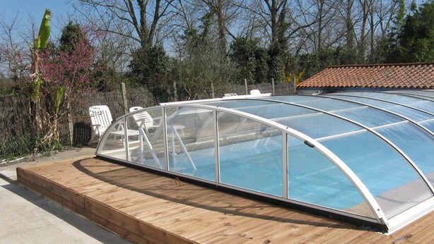A glass-enclosed swimming pool with a wooden deck and a white chair. - Longère vendéenne (Saint Hilaire de riez)