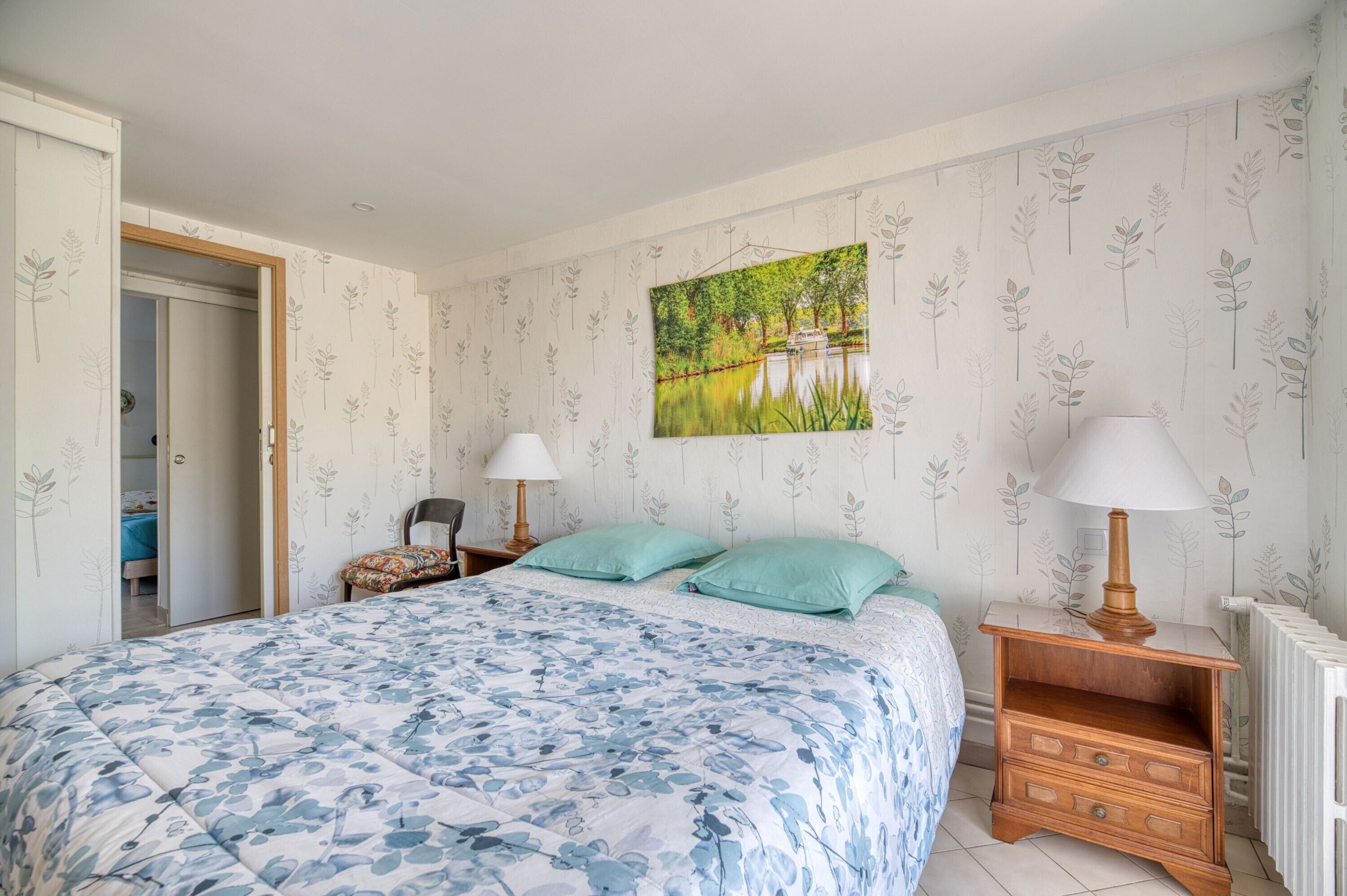 A bedroom with a bed, two bedside tables with lamps, a wall-mounted painting, and a radiator.