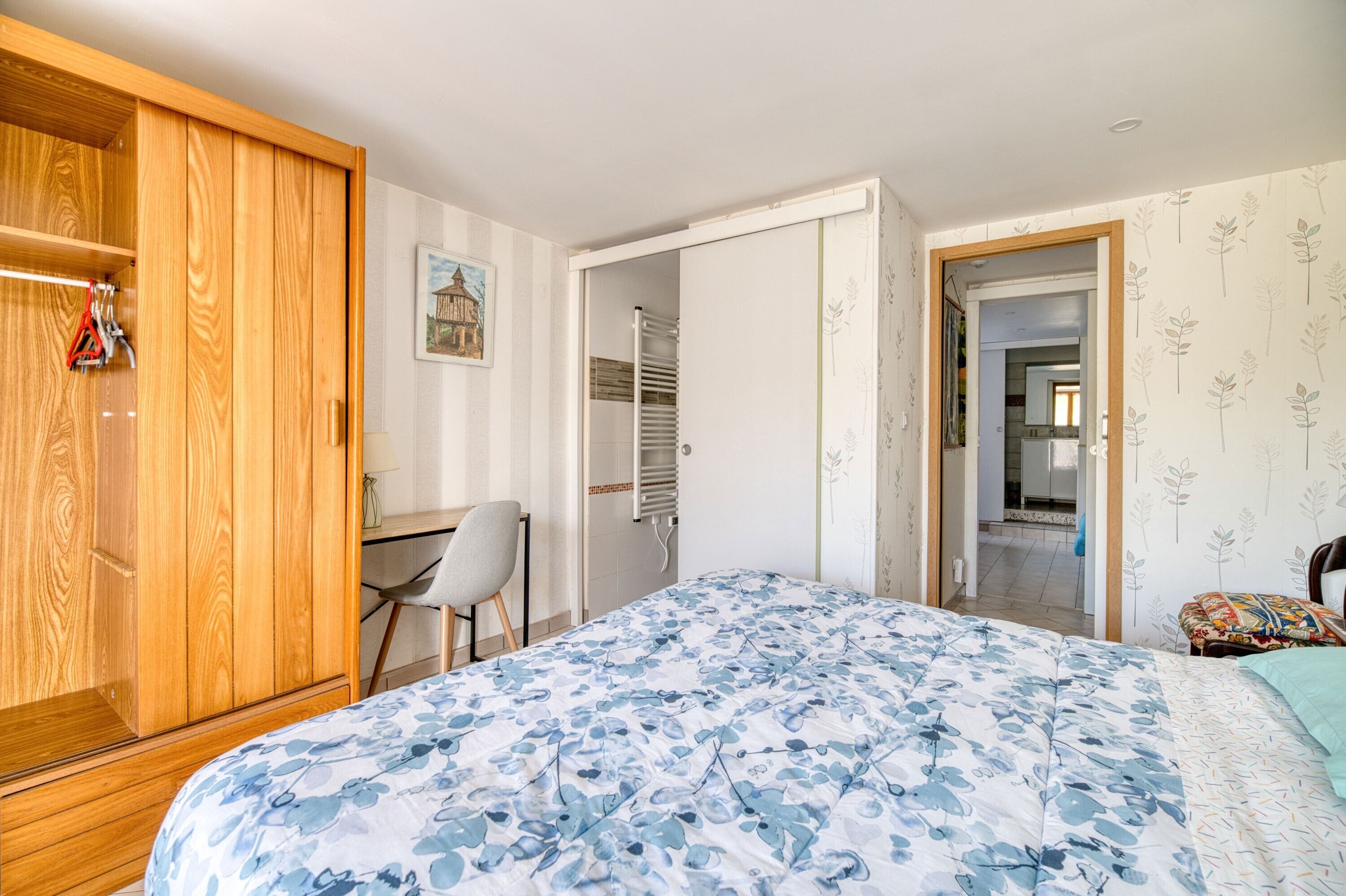 A bedroom with a bed, a wooden wardrobe, a desk with a chair, and a bathroom visible through an open door.