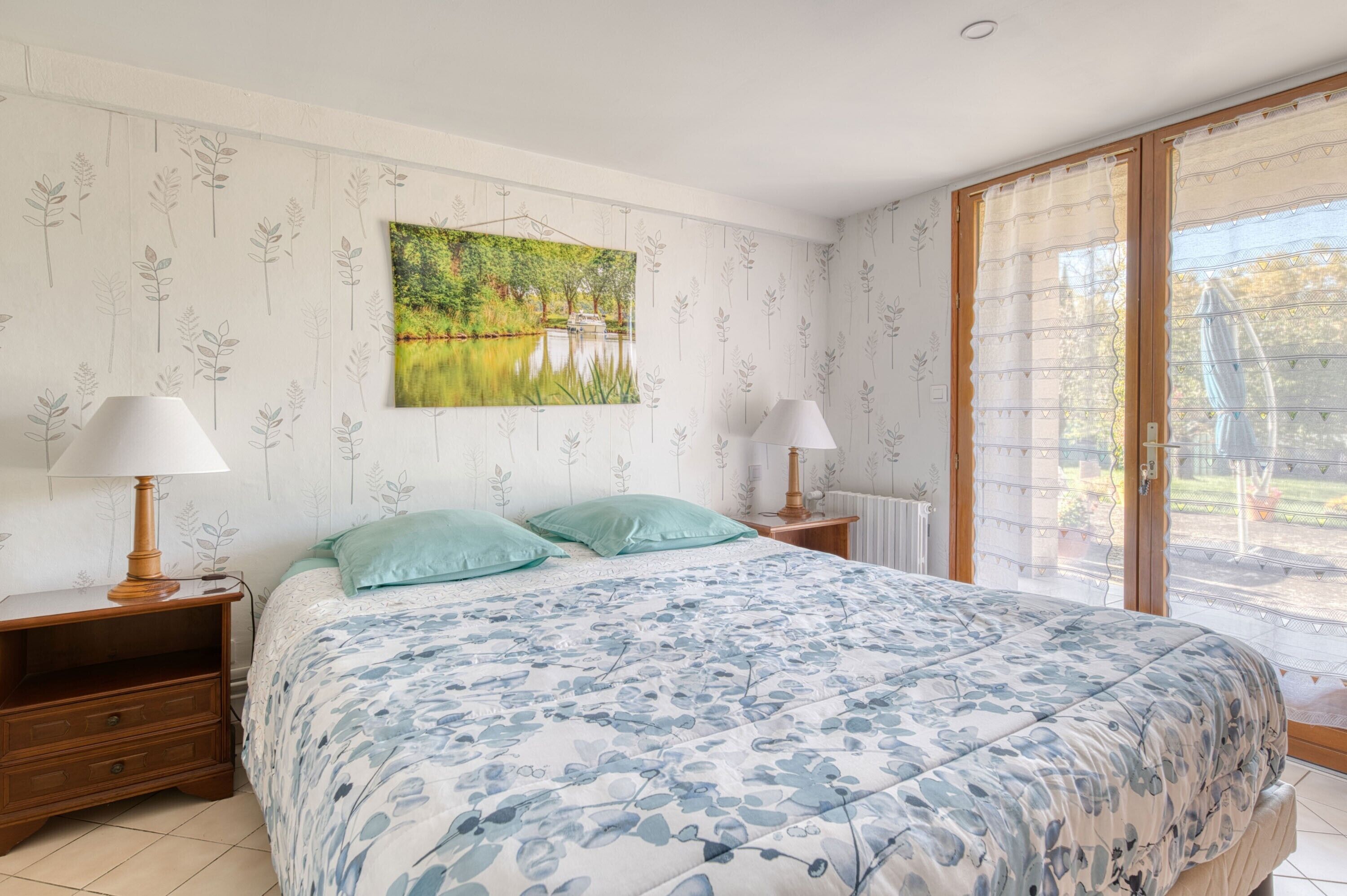 Un dormitorio con cama, dos mesitas de noche, ventana con vistas y un cuadro en la pared.