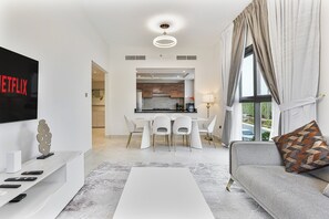 A modern living room with a white sofa, a coffee table, and a TV mounted on the wall. There is a dining area with a white table and chairs, and a kitchen in the background.