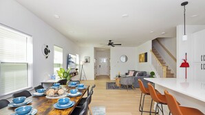 Un espace de salle à manger moderne avec une table en bois dressée pour six personnes, entourée de chaises noires. Un escalier mène à un niveau supérieur, et un balai rouge est accroché au mur.