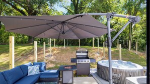 A backyard with a barbecue grill, a hot tub, and a blue sofa under a canopy. - Uptown Retreat Sleeps 10 w/Hot Tub, Games /Fenced (Charlotte)