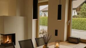 A modern dining room with a wooden table, black chairs, a vase with dried flowers, a lit candle, a fireplace, and a view of the outdoors.