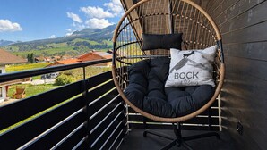 A wicker chair with a cushion and a pillow on a balcony with a view of a village and mountains.