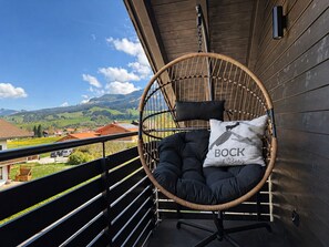 A wicker chair with a cushion and a pillow on a balcony with a view of a village and mountains.