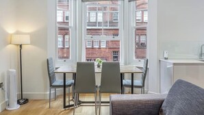Habitación de esquina con una mesa de comedor, dos sillas, una lámpara y vistas a edificios de ladrillo rojo.