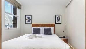 Una cama bien tendida con sábanas blancas y una pila de toallas dobladas, un cabecero de madera y dos cuadros en la pared.