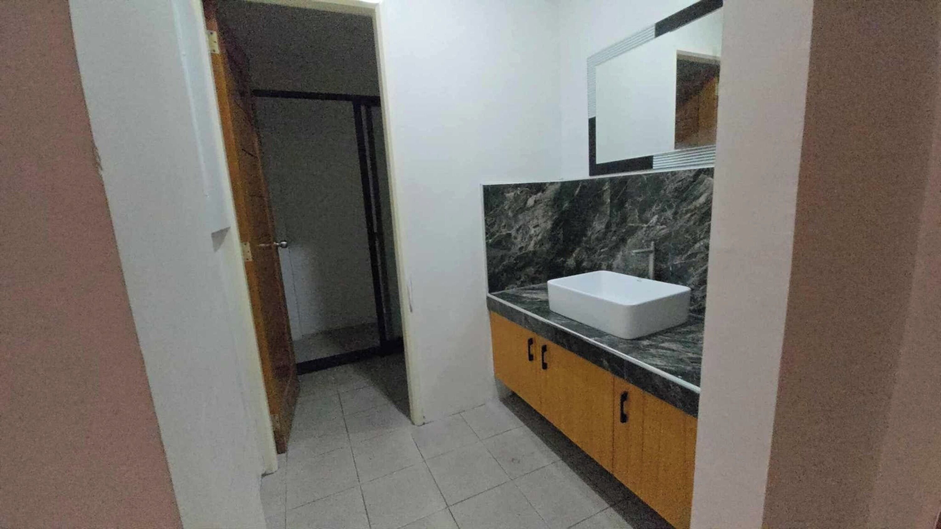 Un baño con lavamanos blanco, encimera de mármol y espejo encima. Se ve una puerta que da a otra habitación.