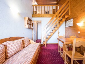 Una habitación acogedora con una escalera de madera, una cama con ropa de cama escocesa, una mesa de comedor con sillas y un pequeño estante de madera.