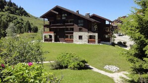 A large, multi-story wooden building with stone accents, surrounded by lush greenery and a clear sky.