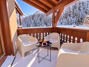 A covered outdoor seating area with two white chairs and a small table, surrounded by a wooden fence, overlooking a snowy landscape with trees.