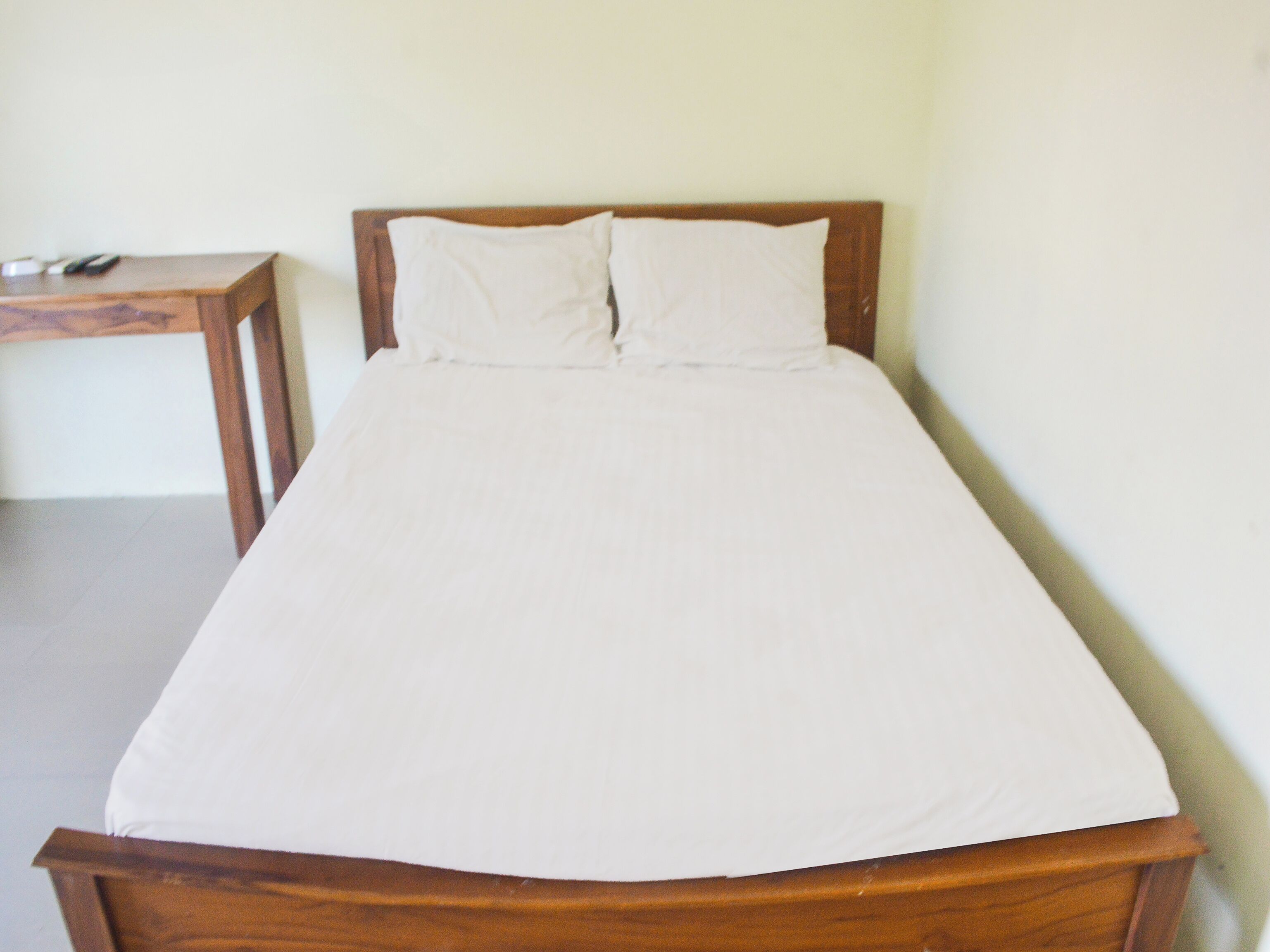 Eenpersoonsbed met houten hoofdbord en witte beddengoed in een kamer met een effen muur en een klein houten tafeltje ernaast.