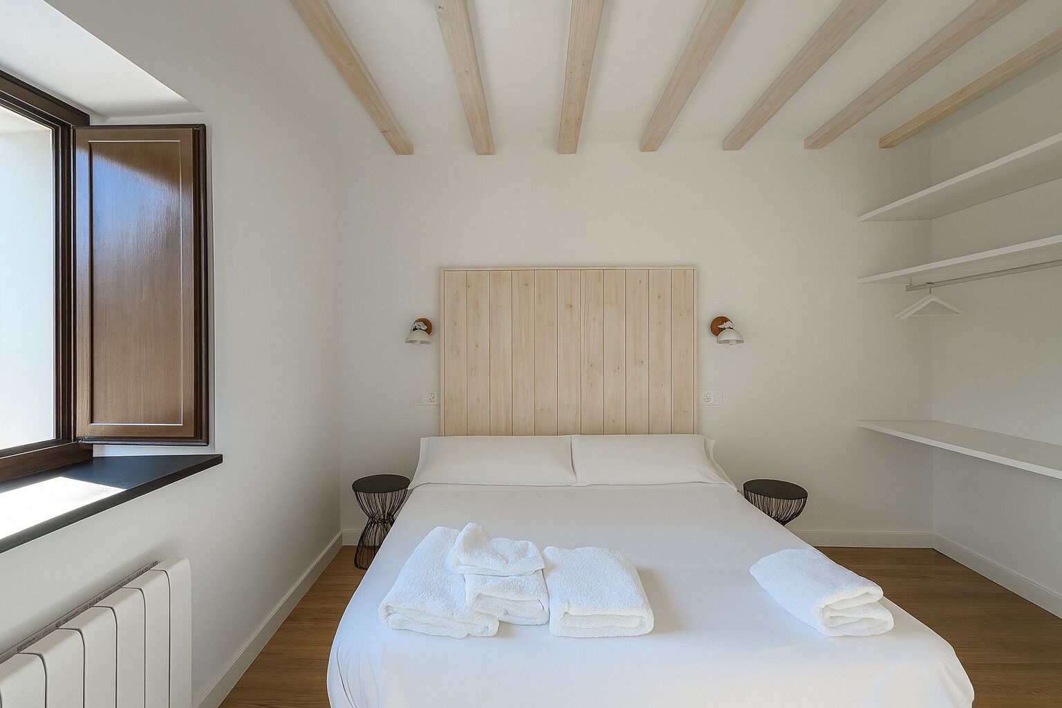 Une chambre à coucher avec une tête de lit en bois, un lit recouvert de draps blancs, deux tables de chevet, une fenêtre et un radiateur.