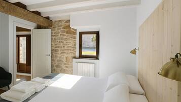 A bedroom with a stone wall, a bed with white linens, a wooden ceiling, and a window with a view.